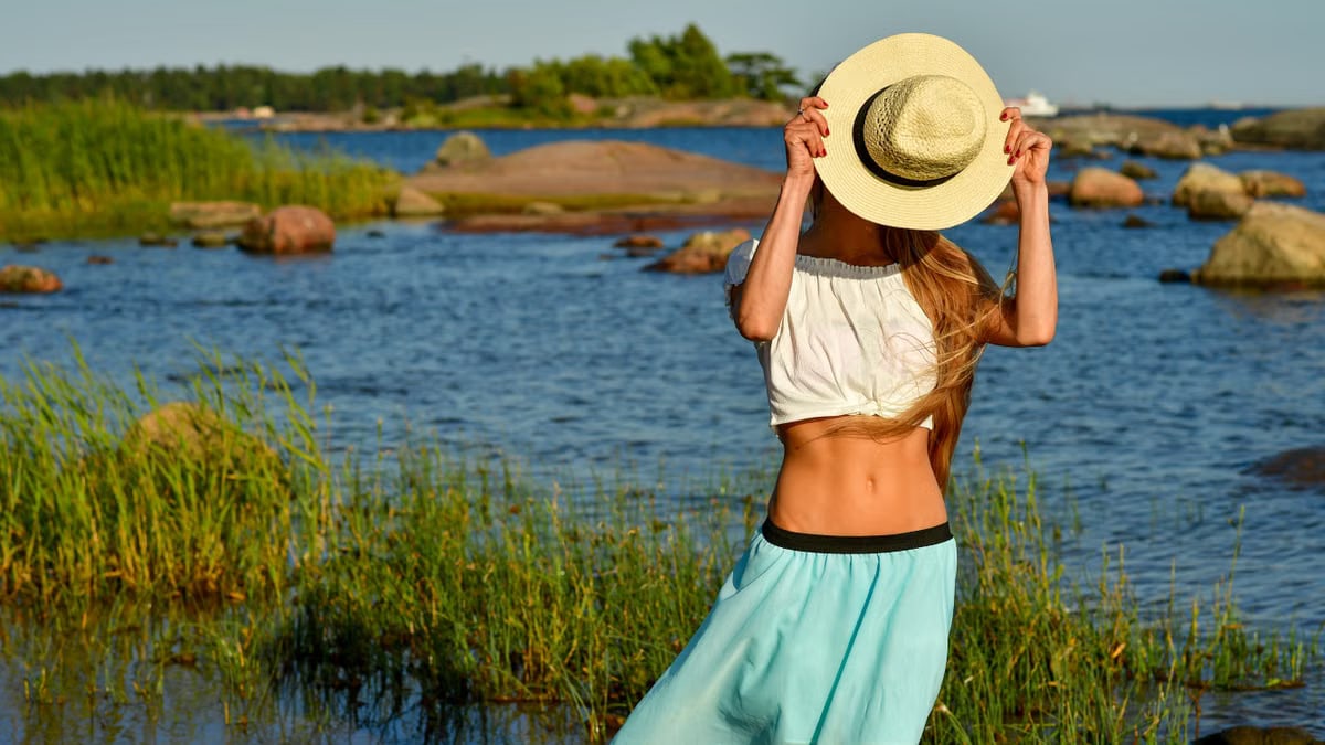 Woman enjoys a summer day by the rocky beach in Helsinki, hiding face with a straw hat